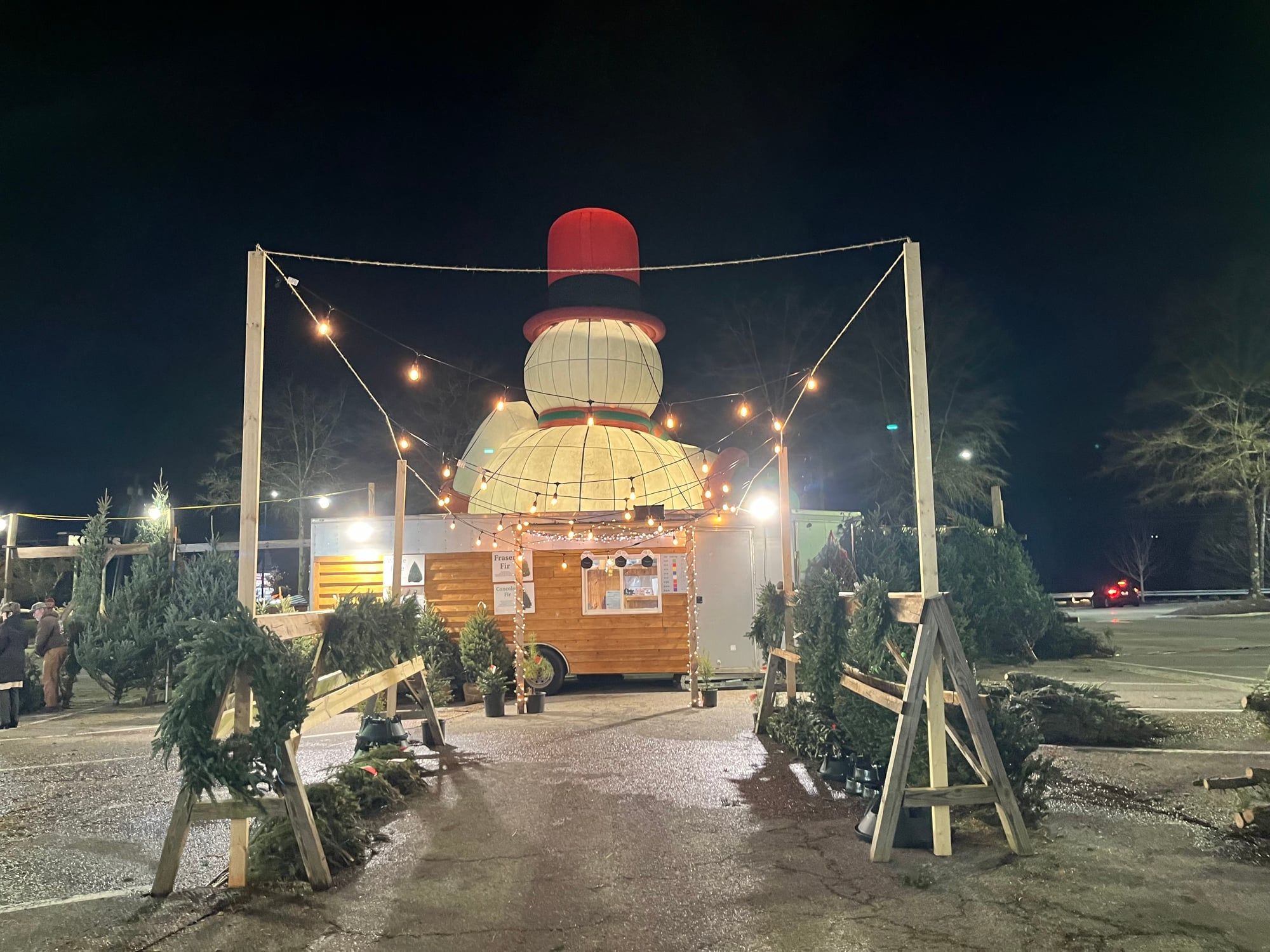 The Roanoke lot at night: a lit timber entrance strung with bulbs, wreaths on the rails, the cedar sales trailer and Frosty glowing behind it