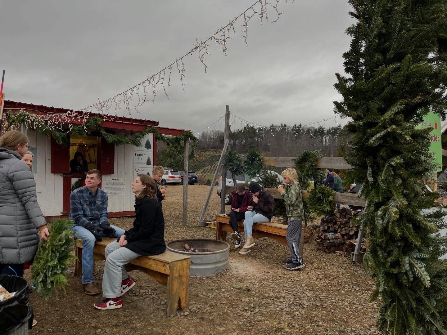 Customers on benches holding fresh wreaths under string lights beside the wreath shed